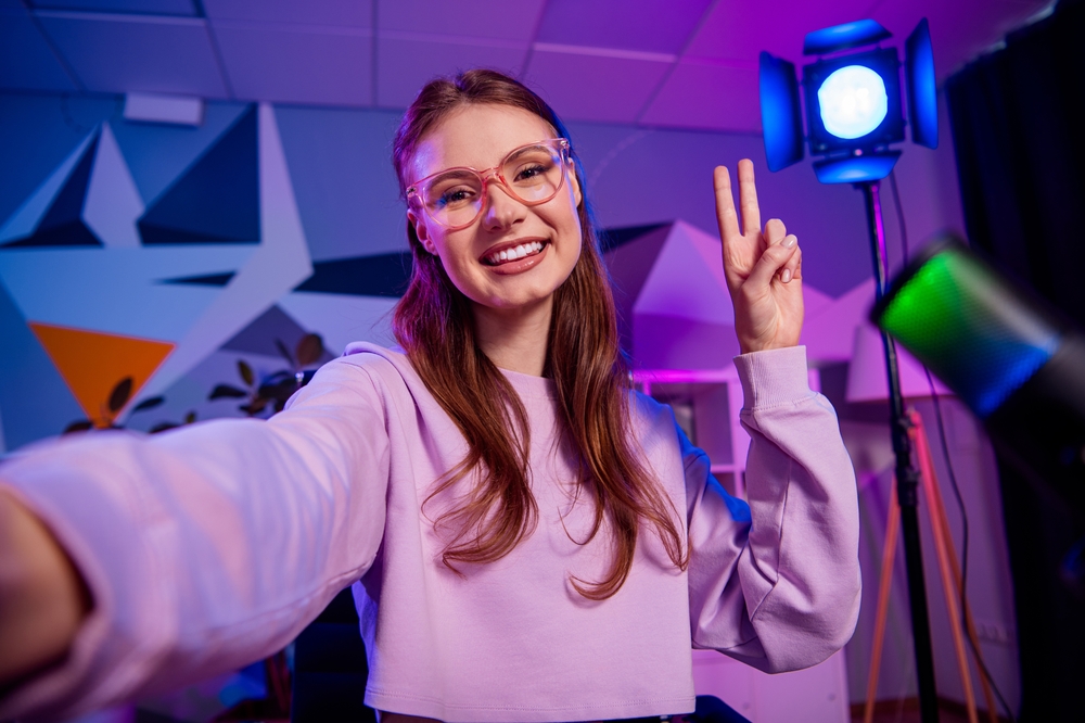 A cheerful young female content creator takes a selfie in a vibrant studio setting with neon lighting, highlighting her passion for media, blogging, and digital influence.