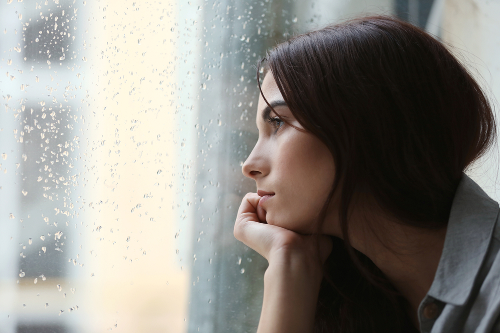 Depressed young woman near window at home, closeup
