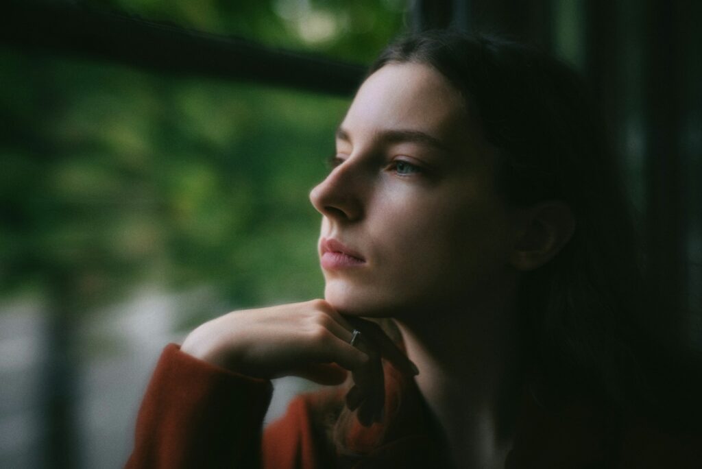 A woman looking out a window with her hand on her chin
