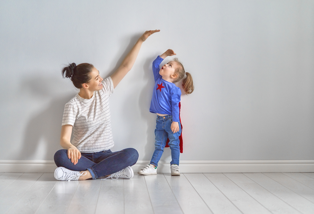 Mother is measuring growth of child daughter near empty wall. Girl in superhero costume.