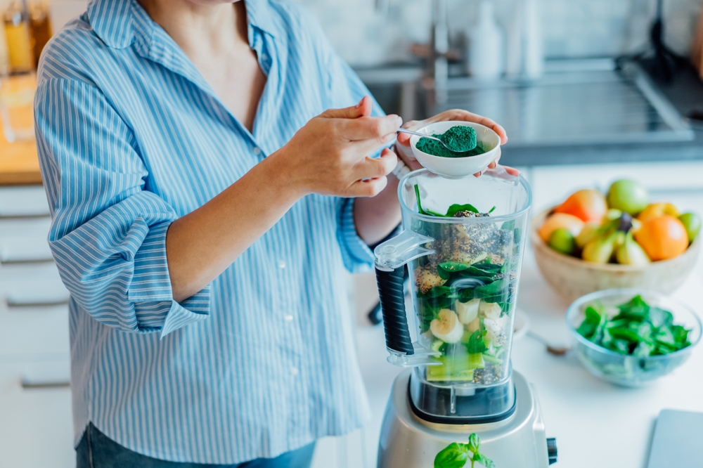 Woman adding spirulina green powder during making green smoothie on the kitchen. Superfood supplement. Healthy detox vegan diet. Healthy dieting eating, weight loss program. Selective focus.