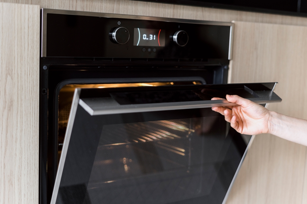 Cropped view of woman hand open glass door on electric oven, cooking food at home. Concept of household appliance, baking equipment at kitchen. Female using microwave to heat meal