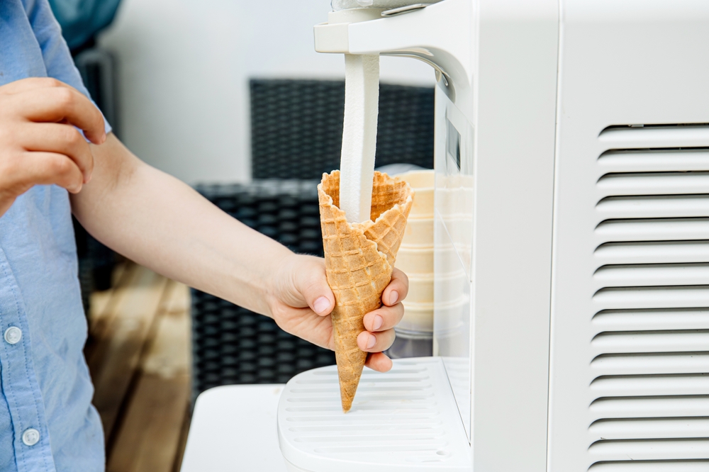 Close up view of child hand using home use ice cream maker machine. Making soft serve ice cream and pouring in cone outdoors in garden yard, summer birthday party.