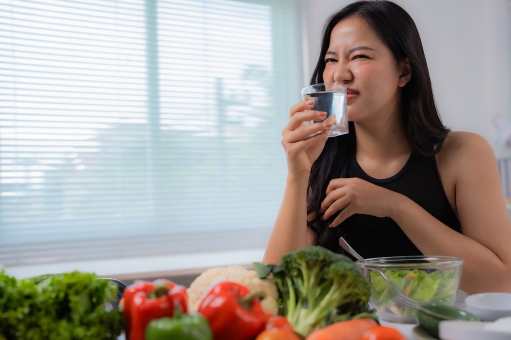 Young woman is drinking a glass of water while making a disgusted expression. Sitting at the kitchen table with a lot of fresh vegetables. Including broccoli. Bell peppers. Lettuce. And cauliflower