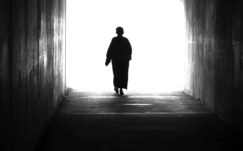 A black and white high contrast silhouette of figure in a landscape of a single lady or woman stepping or walking through a tunnel into and twowards a bright light. 