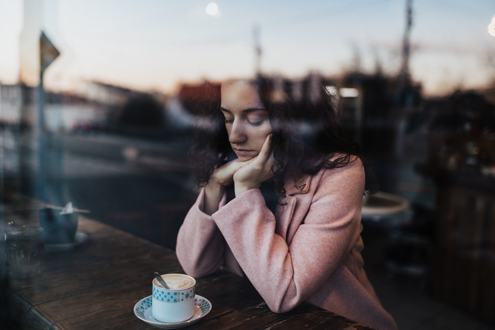 Sad,pensive young woman drinking coffee and looking out of the cafe window.