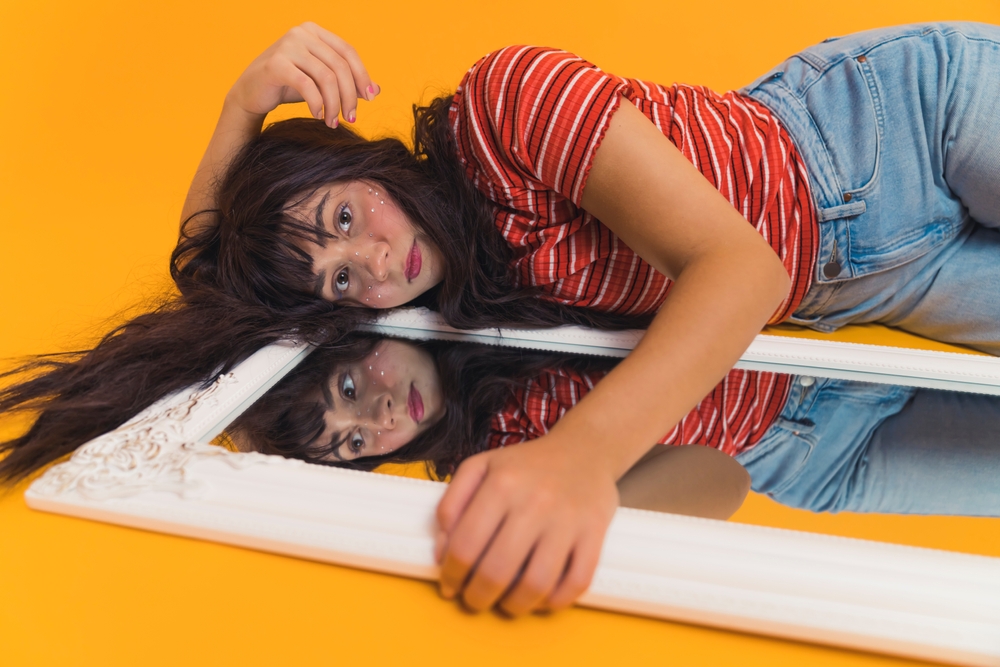 Teenagers with psychological problems such as body dysmorphia. Sad long-haired female caucasian teenager in stripped t-shirt lying next to bedroom mirror and touching in with her hand. Studio shot