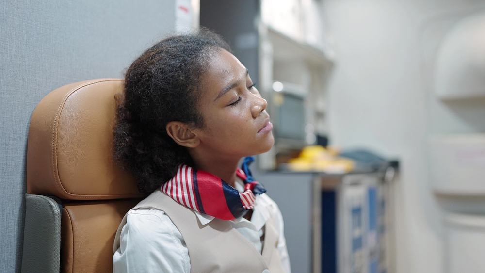 Woman african american stewardess, flight attendant sitting on seat in airplane looking tired after a long international flight itinerary. Tired flight attendants resting between flights