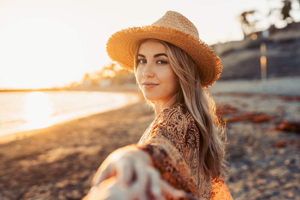 One cute girl walking on the sand of the beach holding her boyfriend’s hand. Enjoying travel and vacation summer lifestyle together outdoors at sunset.