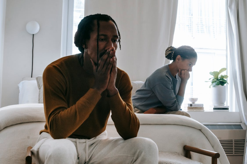 ethnic couple sitting on bed in sunlit room making thinking gestures