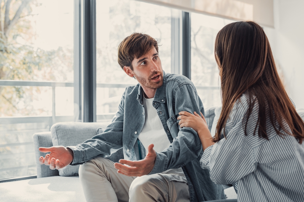 Attractive man is depressed, sitting on the couch ignoring woman, having a dispute with wife or boyfriend, looking desperate and hopeless, having unresolved problem or question