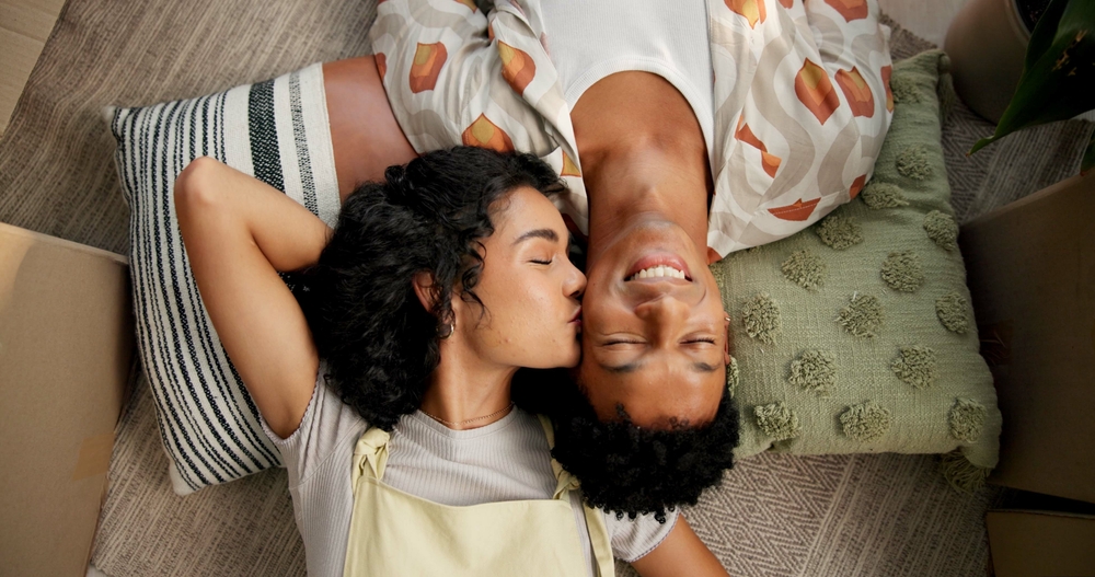 Couple, above and kiss on cheek or new home floor, happiness and love for relationship or moving boxes. Relax, property investment and affection in apartment living room, people care and homeowners