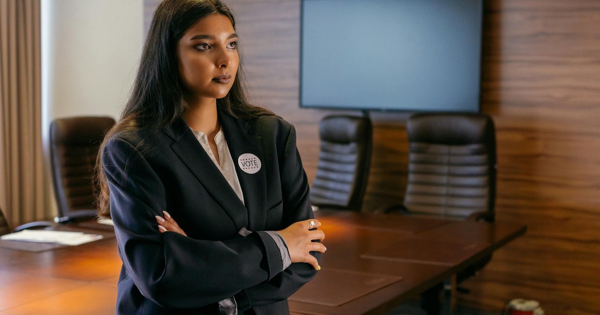 A confident woman in a business suit stands with arms crossed in a corporate boardroom.