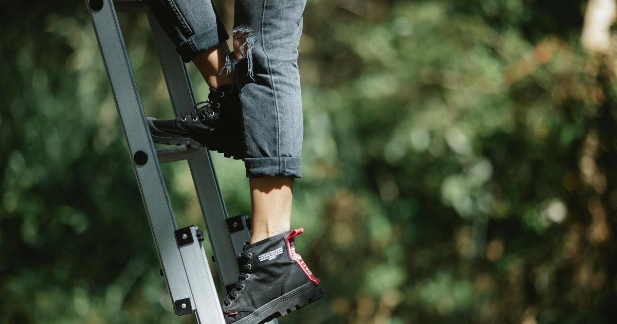 Side view of crop unrecognizable person in stylish boots and jeans ascending stepladder in green forest