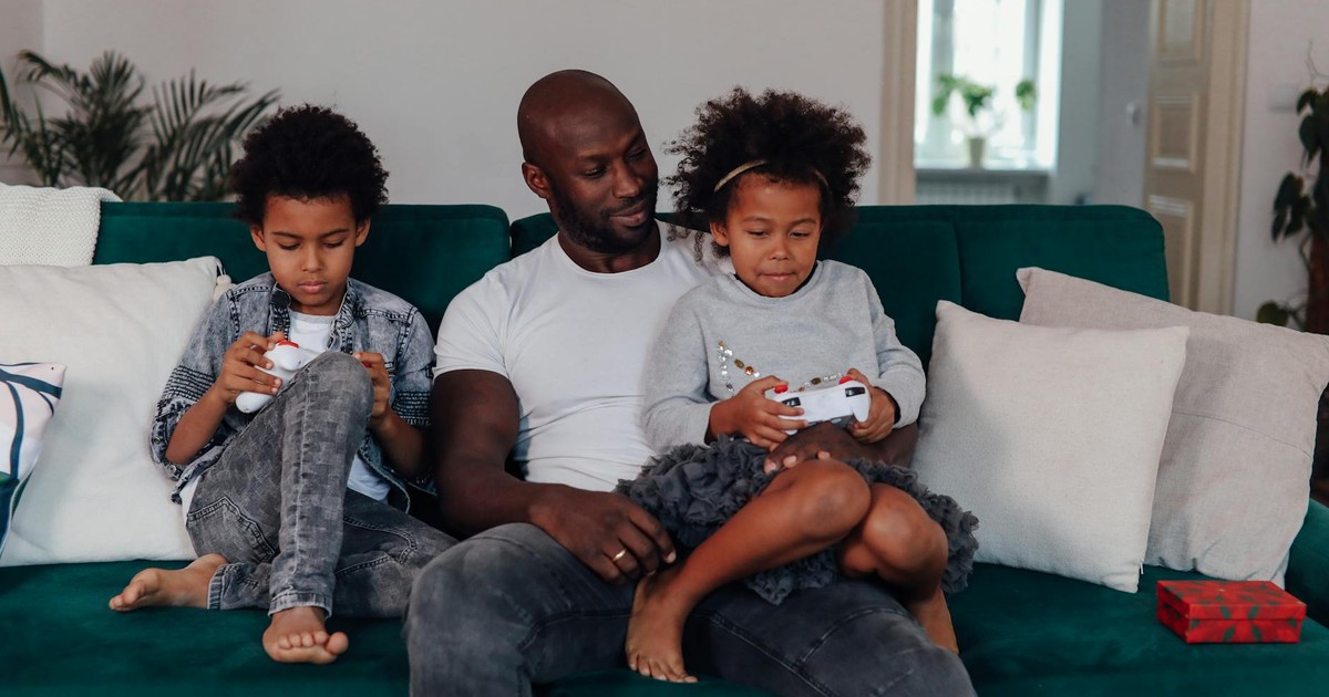 A father and his two children enjoy playing video games together indoors, promoting family togetherness.