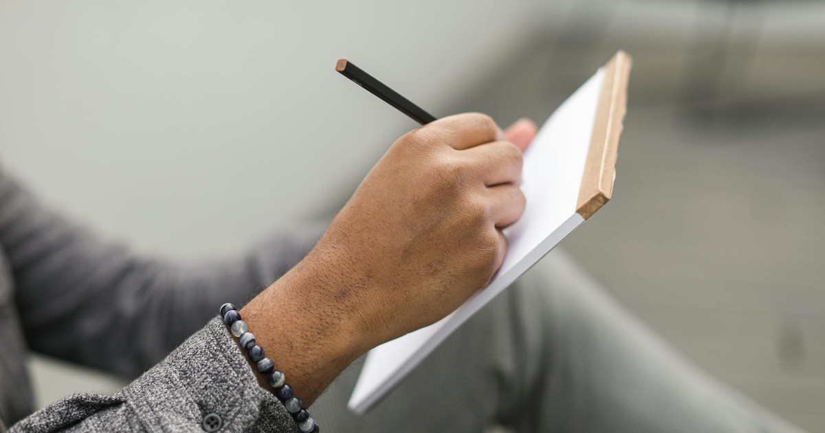 An adult hand writes in a notebook with a pencil, wearing a beaded bracelet and gray plaid sleeves.