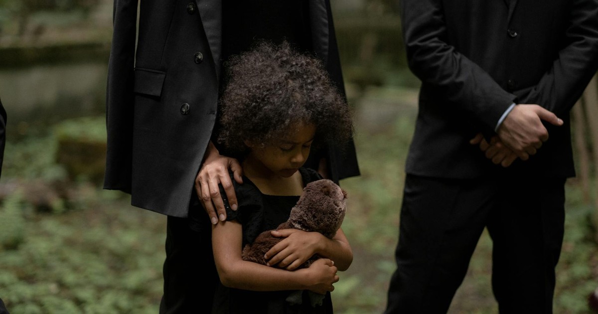 A child holds a plush toy during a somber outdoor funeral service.