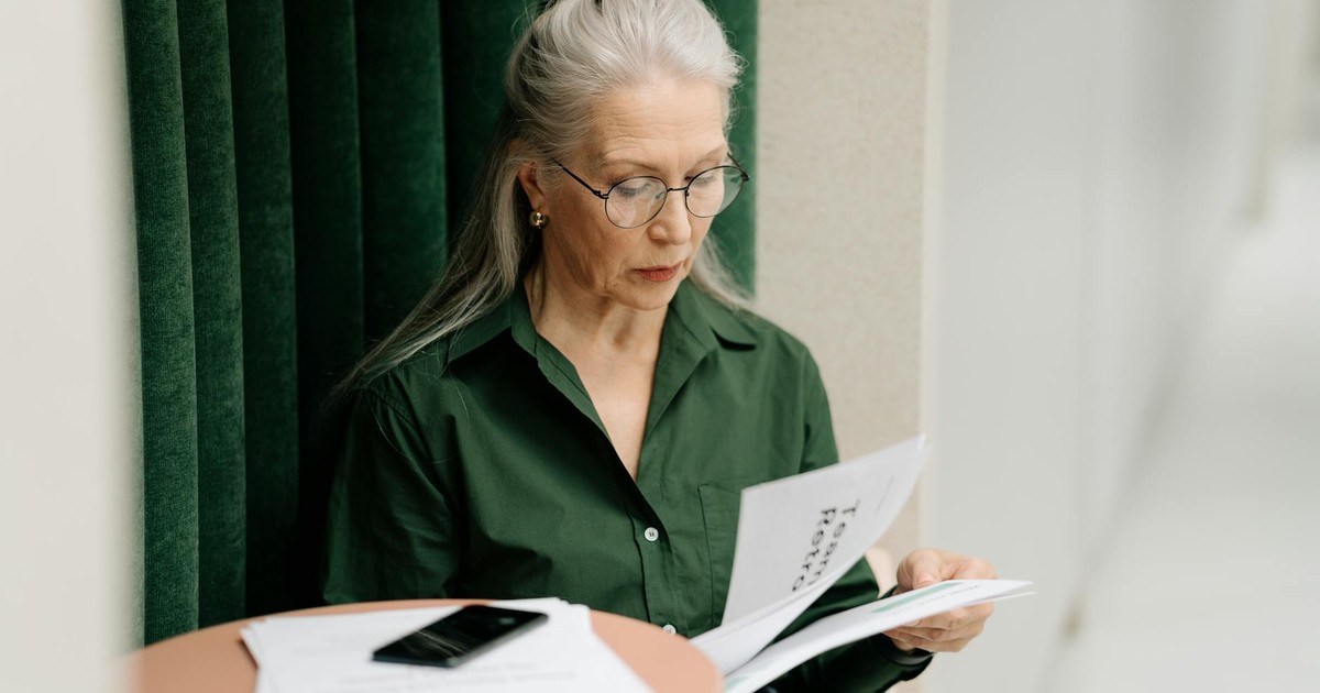 Elderly woman with gray hair reading papers at a small table indoors, focused and thoughtful.