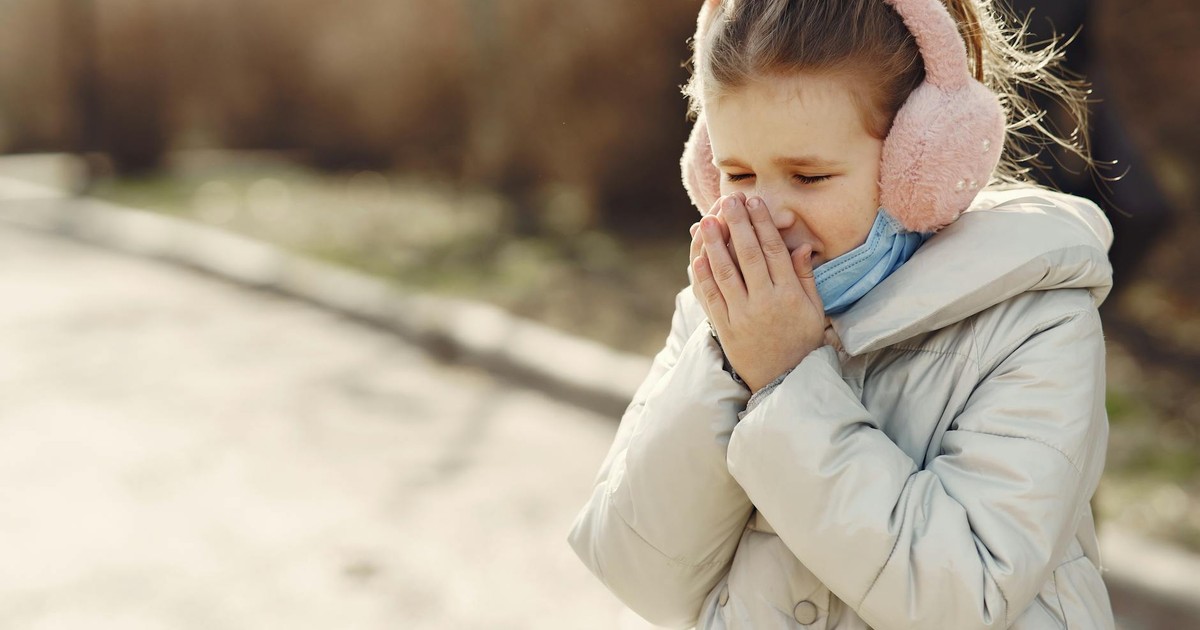 Small child with lowered face mask covering face by hands while sneezing on street