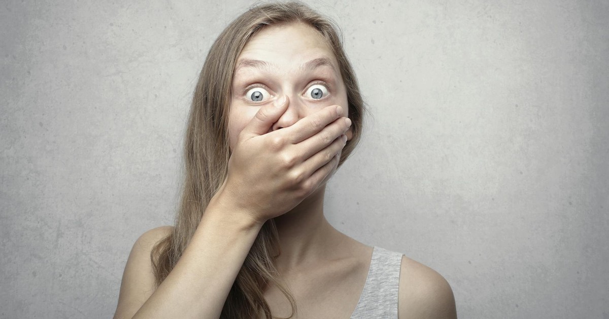 A startled woman covers her mouth expressing surprise against a neutral background.