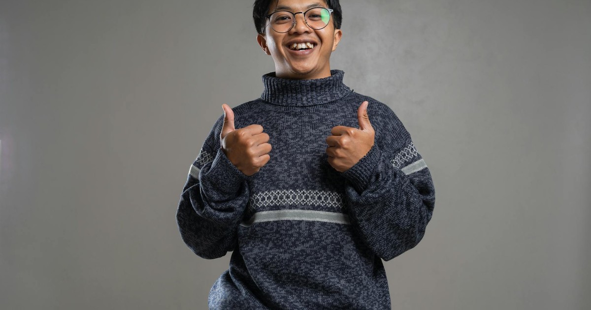 Happy young man wearing eyeglasses and a sweater, giving thumbs up with a big smile indoors.