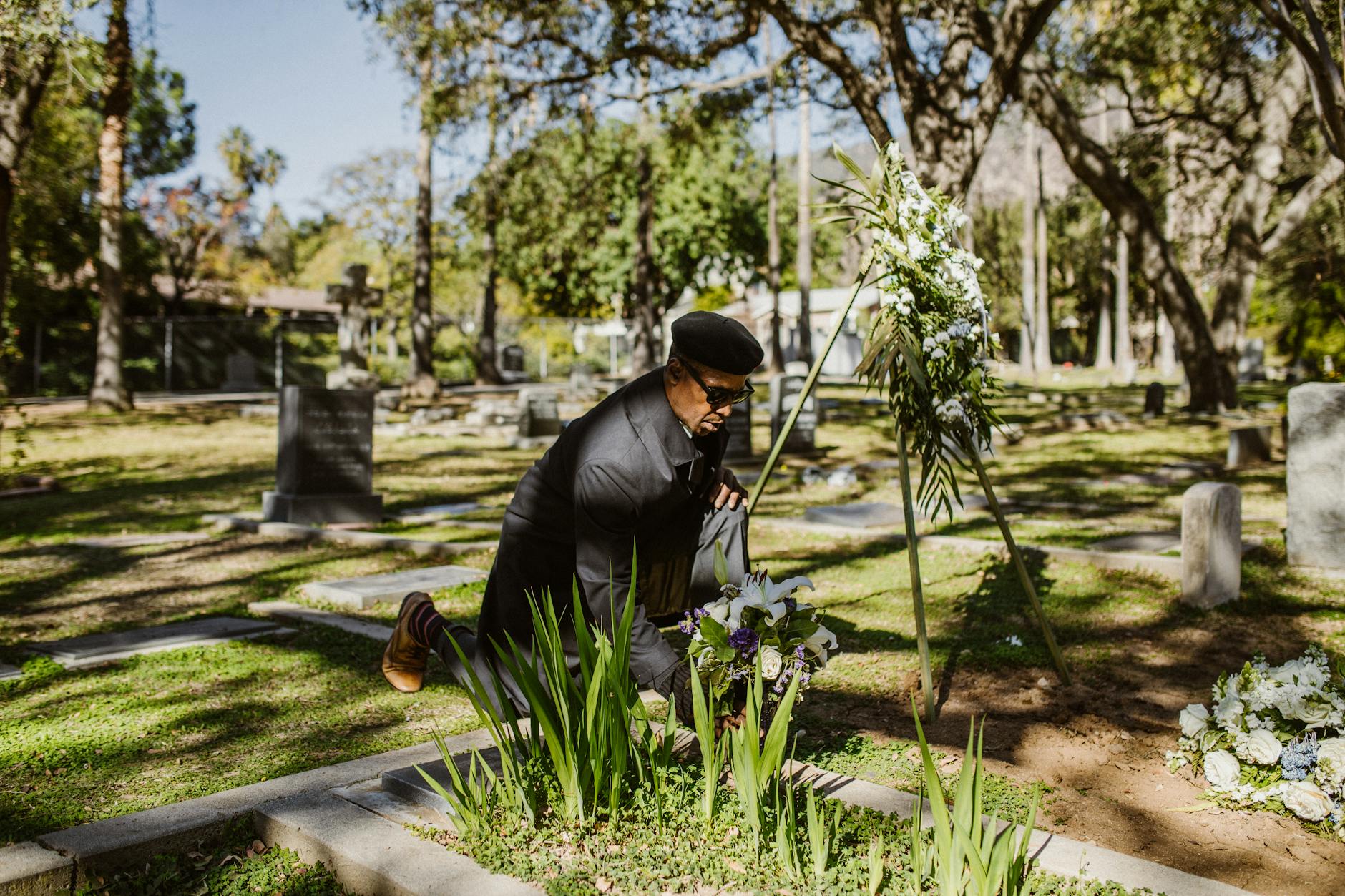 An elderly man in a cemetery mourning with flowers, surrounded by trees.