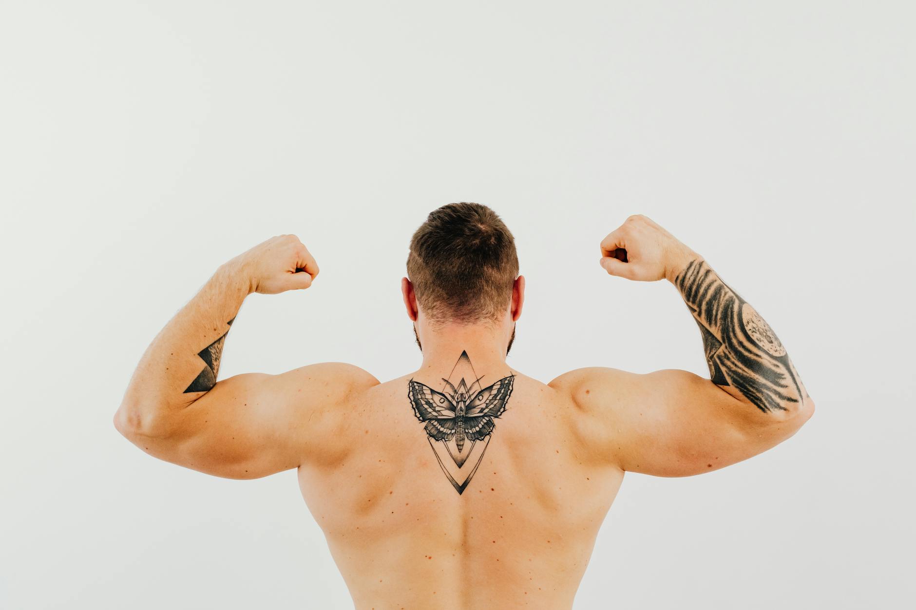 Shirtless man showcasing tattoos and biceps while flexing in a studio setting.