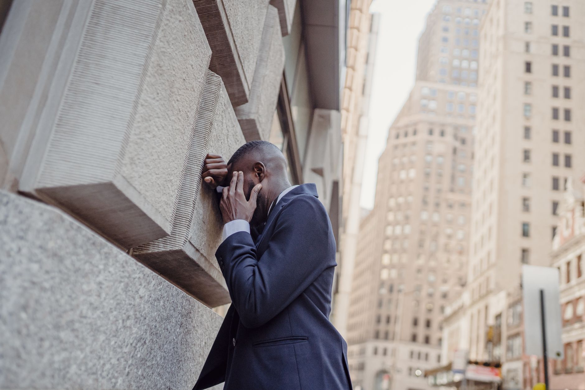 A man in a suit leans against a city wall, seemingly overwhelmed.