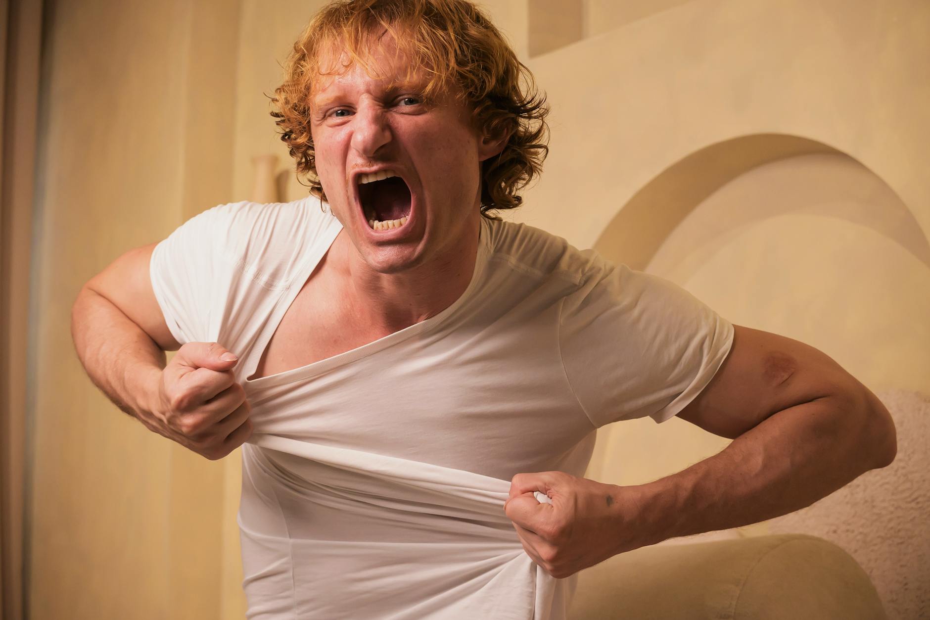 A man exhibiting anger and frustration by tearing his shirt indoors, with an intense expression.