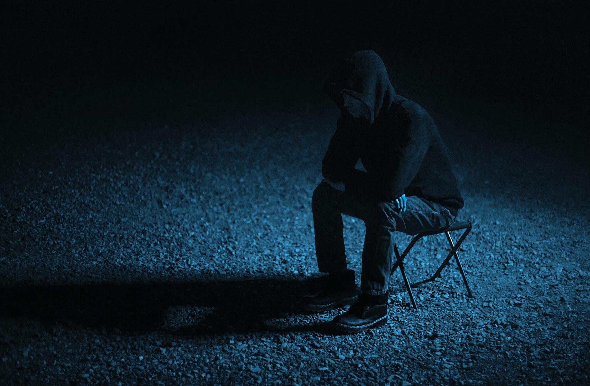 A solitary person in a hoodie sits on a stool under dim lighting, creating a moody night scene.
