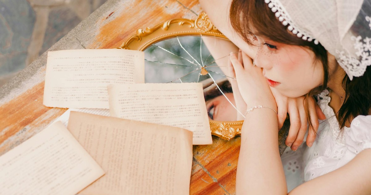 A young woman leans on a table with vintage books and a cracked mirror, reflecting her thoughtful expression.