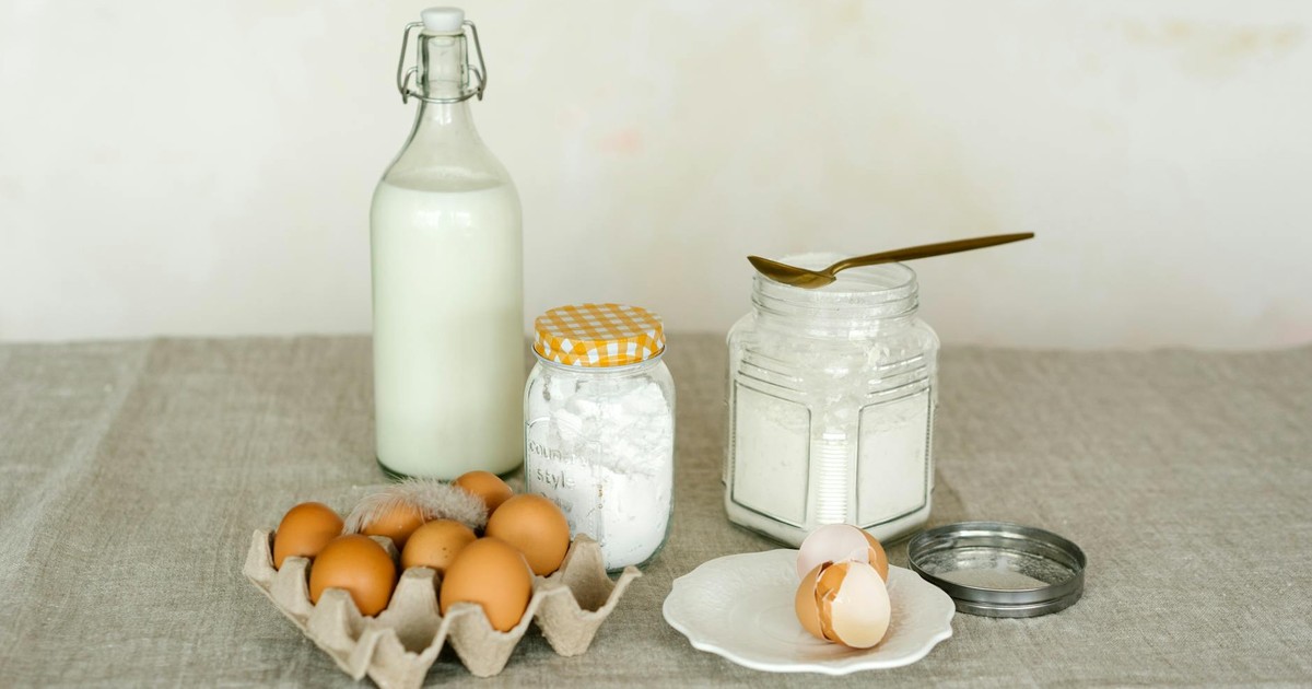 A rustic display of baking ingredients including eggs, milk bottle, and flour on a linen surface.