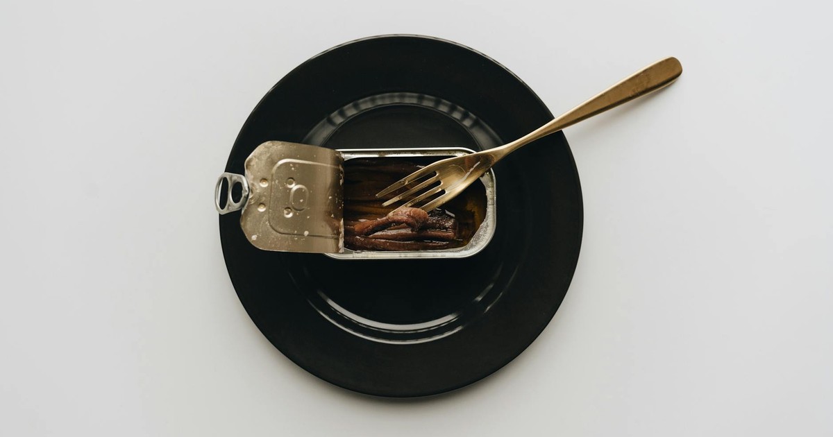 A can of anchovies served on a black plate with a fork in a minimalist setting.
