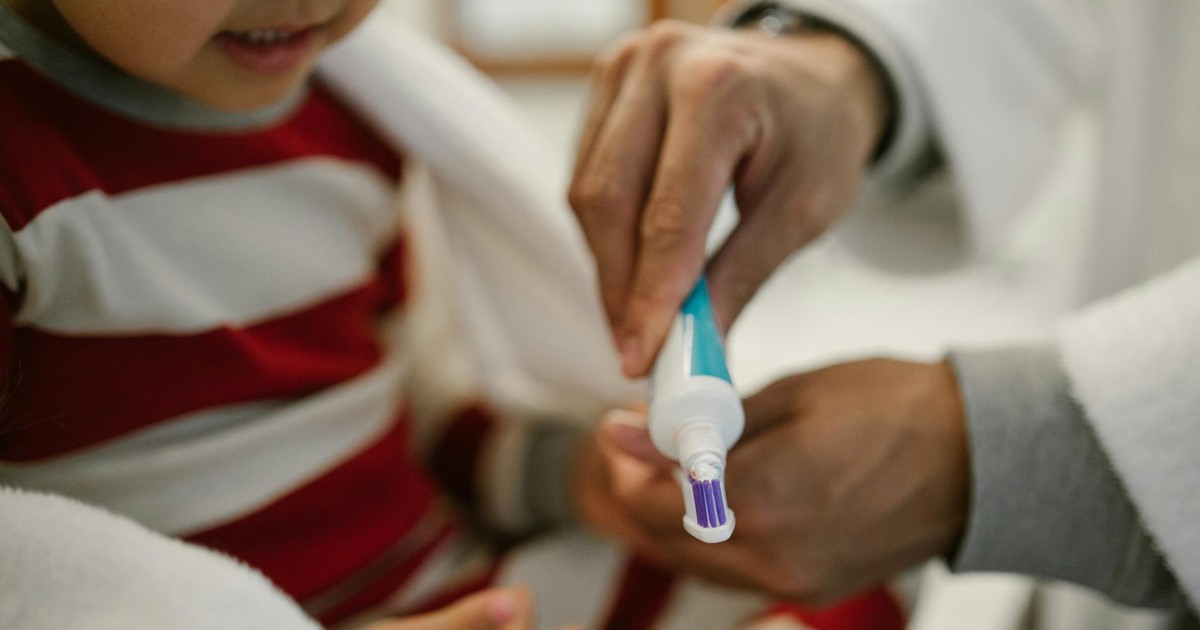 A close-up of a father helping his young daughter apply toothpaste on a toothbrush.