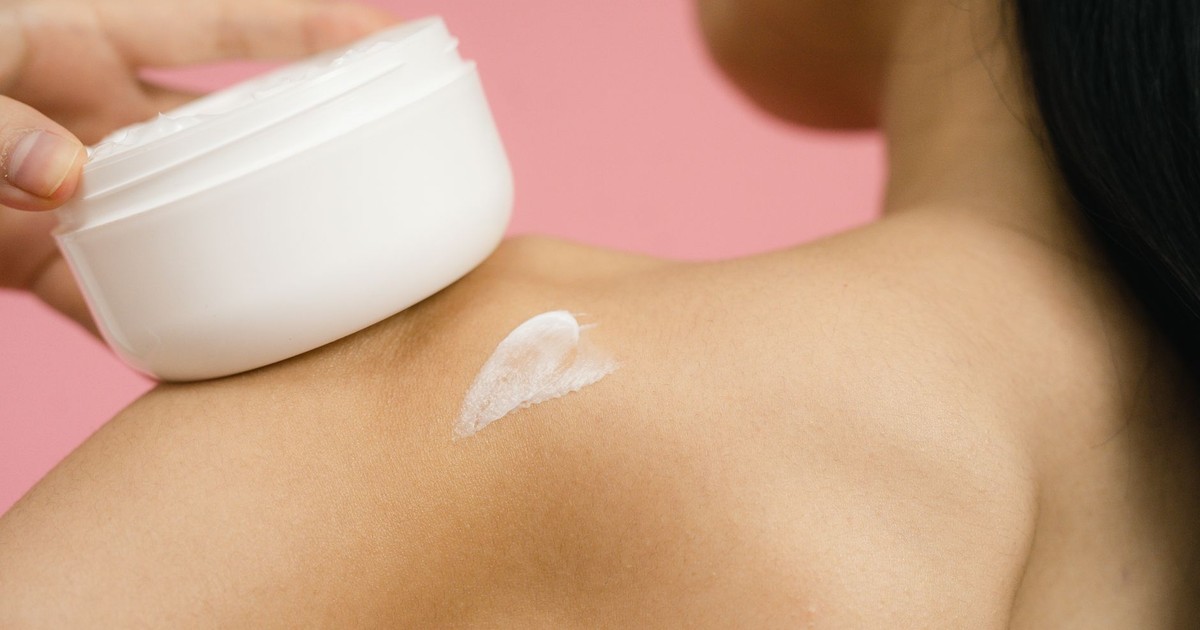 Woman applying skincare cream to shoulder against a pink background.