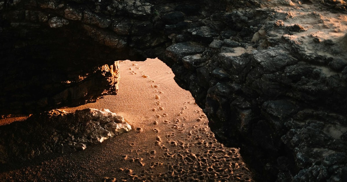 An illuminated coastal cave entrance with footprints in the sand at sunset, creating a dramatic scene.
