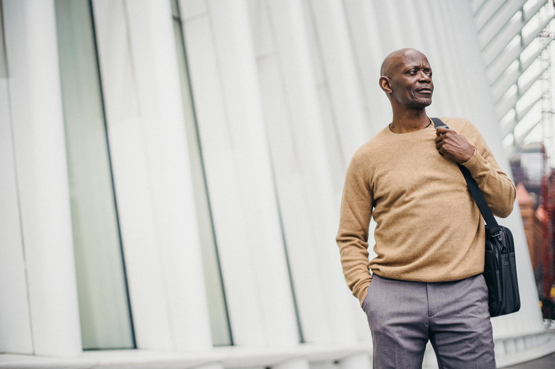 Adult African American bald man in jersey holding bag and standing with hand in pocket looking away on street in center
