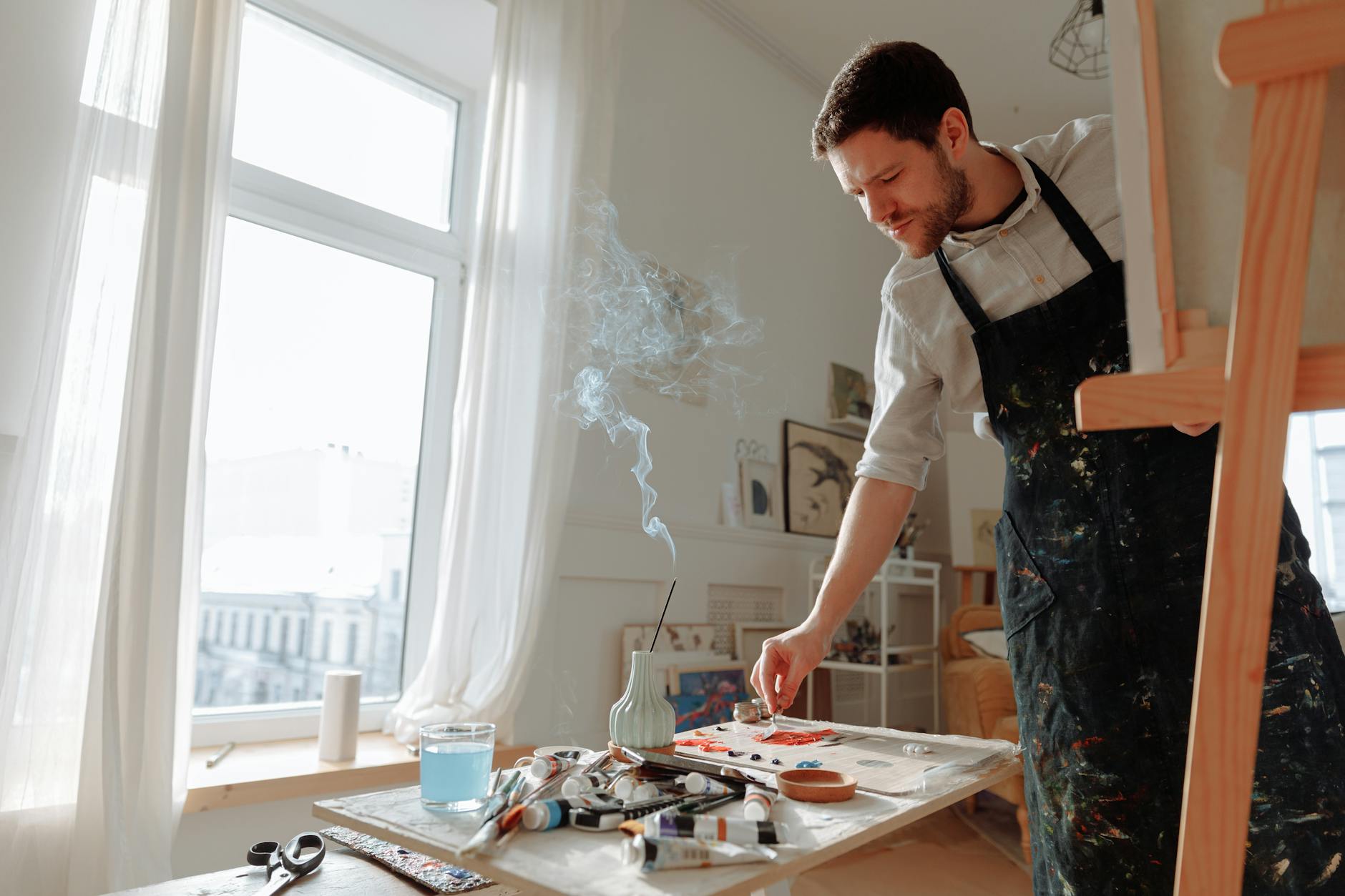 An artist in a sunlit studio works on a painting with focused determination.
