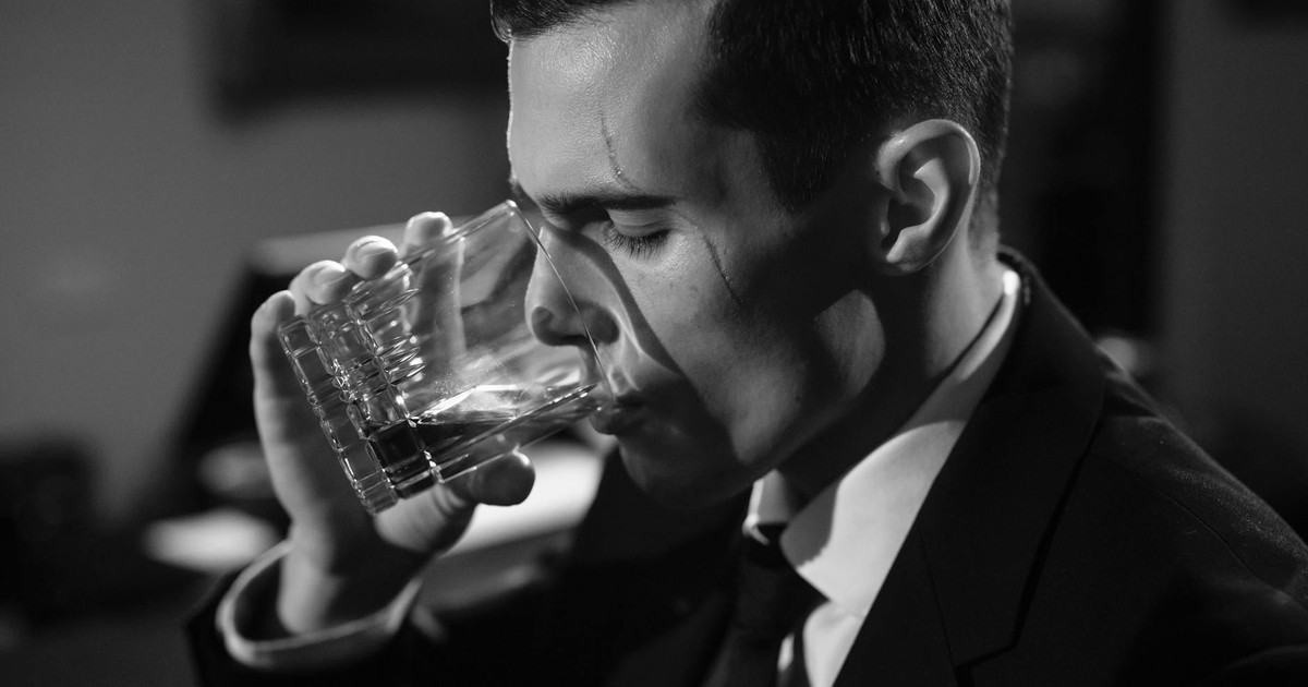 Black and white image of a man sipping whiskey, evoking classic film noir style.