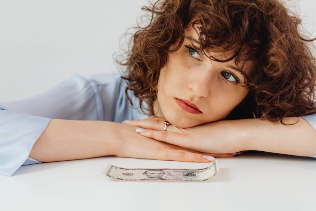 Worried woman with curly hair leaning on white surface with a US dollar bill.

