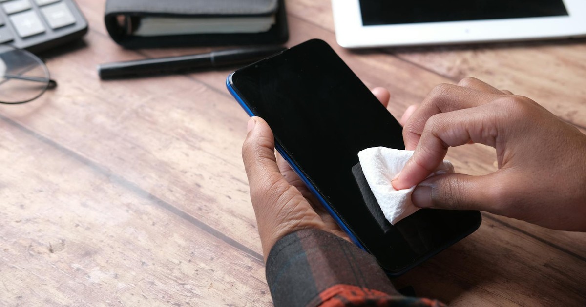 Hands wiping a smartphone with a cloth in an organized workspace. Emphasizes hygiene and device care.