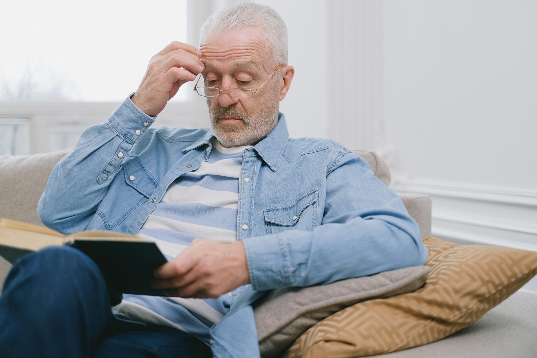 Senior man with glasses reading a book inside a stylish living room.