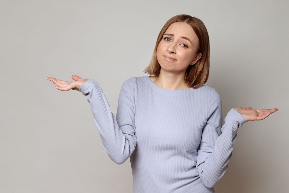 I don't know! Close up studio portrait of shy awkward young woman feeling embarrassed, confused and puzzled, shrugs shoulders, does not know what happened, over grey background.