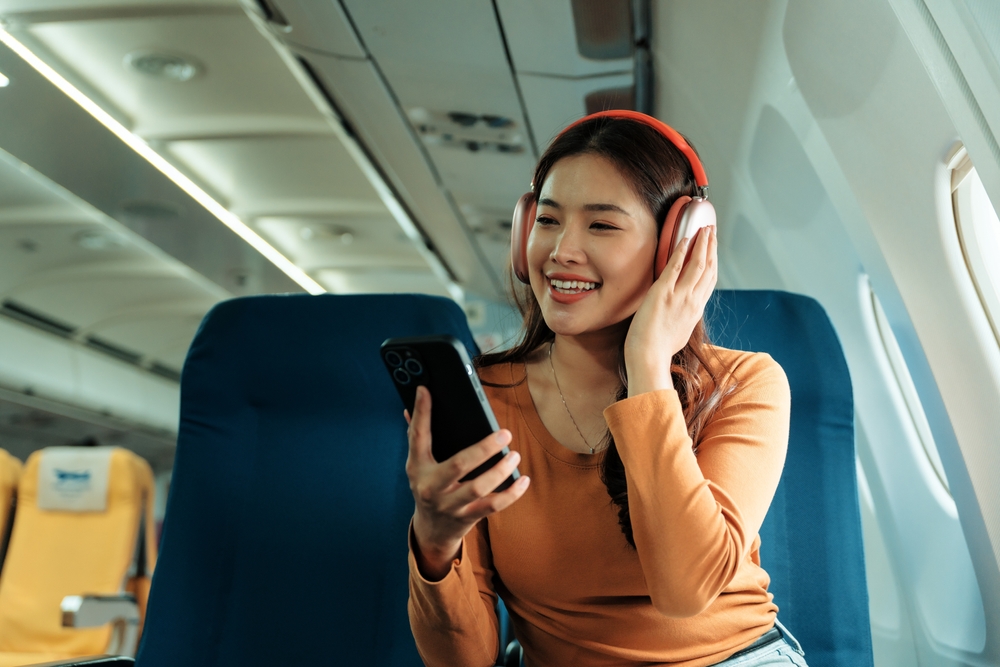 Passenger enjoying entertainment during flight, listening to music with headphones and using smartphone on airplane, smiling happily while relaxing in comfortable seat