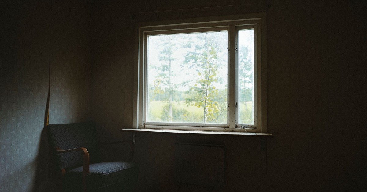 Dark room interior with a chair and window overlooking a tranquil outdoor scene.