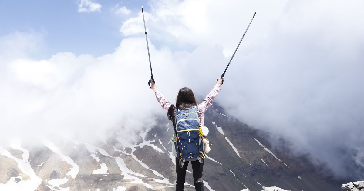 Woman with backpack celebrating reaching a mountain summit with raised hiking poles.