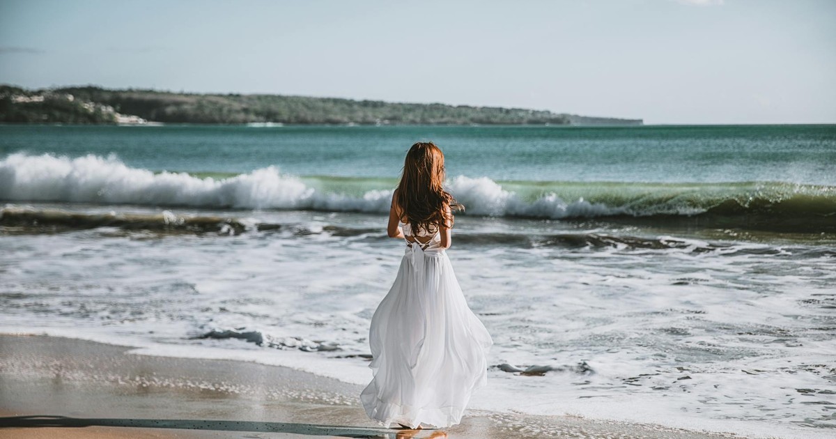 A woman in a white dress strolls along a serene beach with gentle waves under a clear sky.