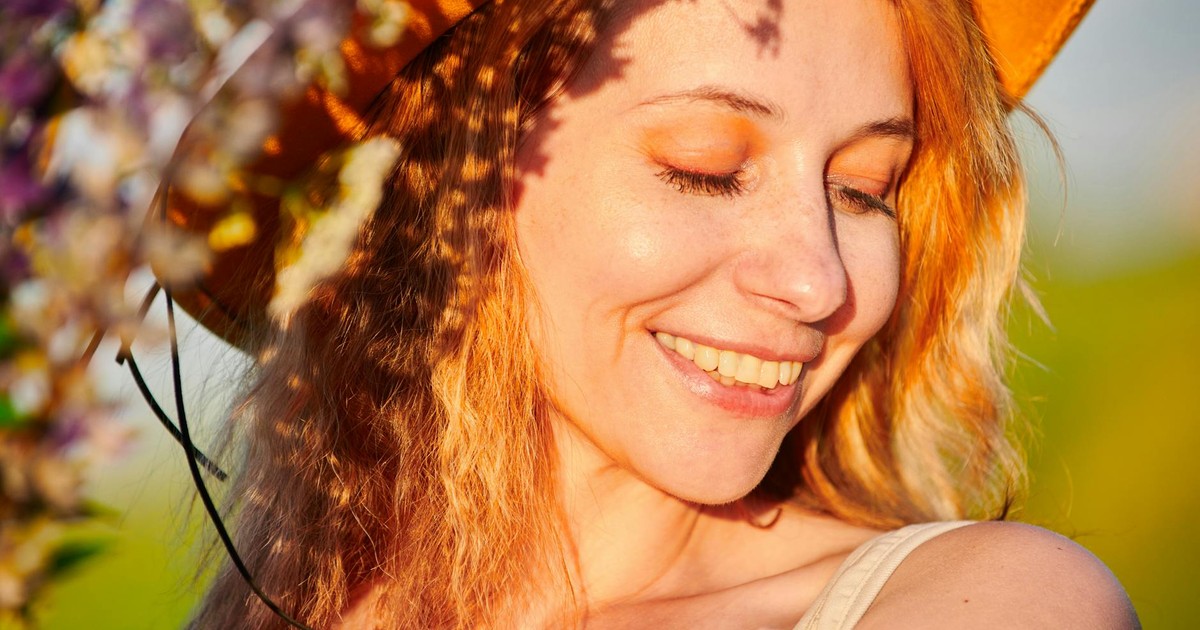 Close-up portrait of a smiling woman in a cowboy hat with vibrant orange hair and makeup, outdoors in sunlight.