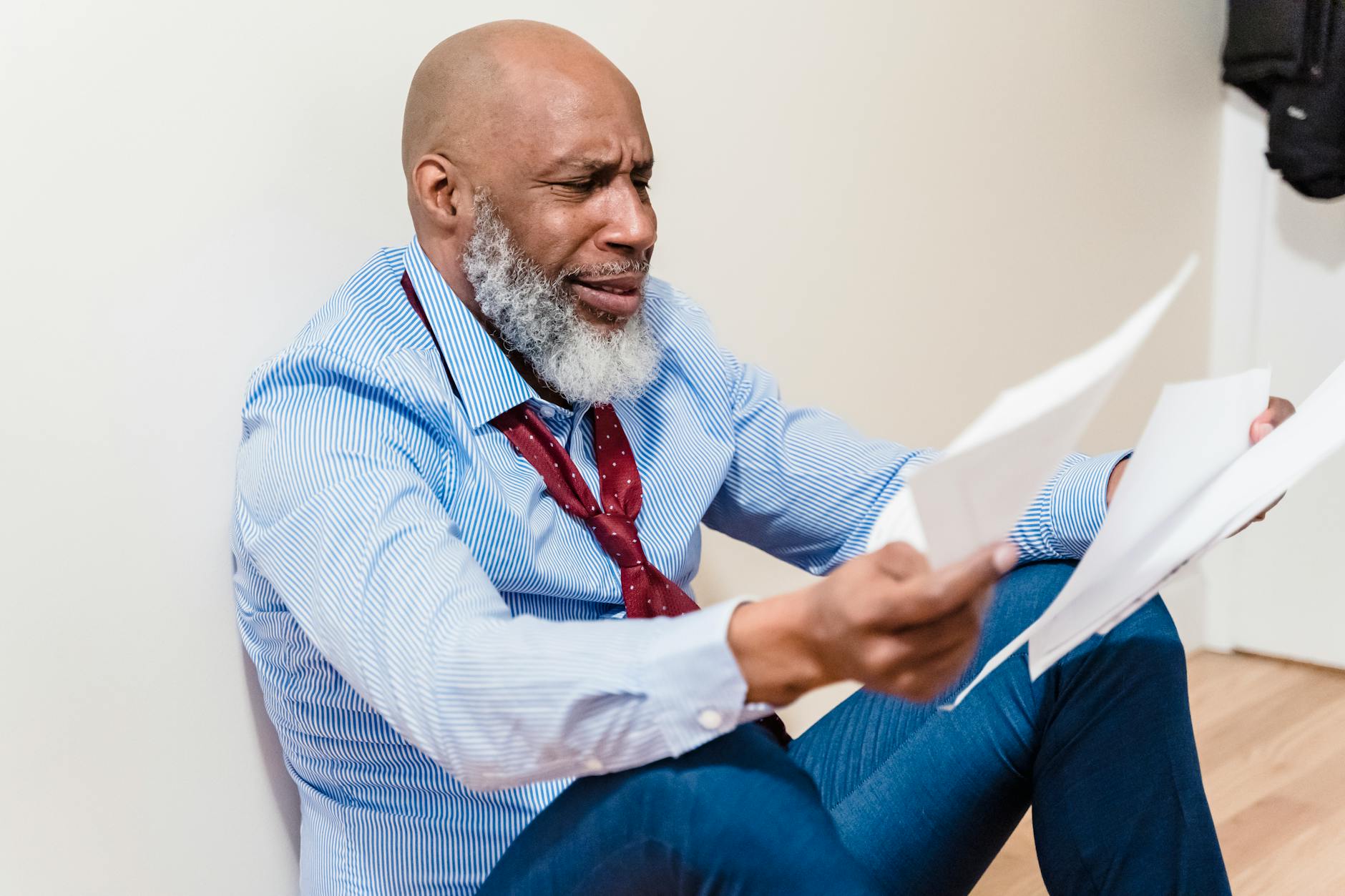 An adult man with a beard looks distressed while sitting on the floor, examining papers.