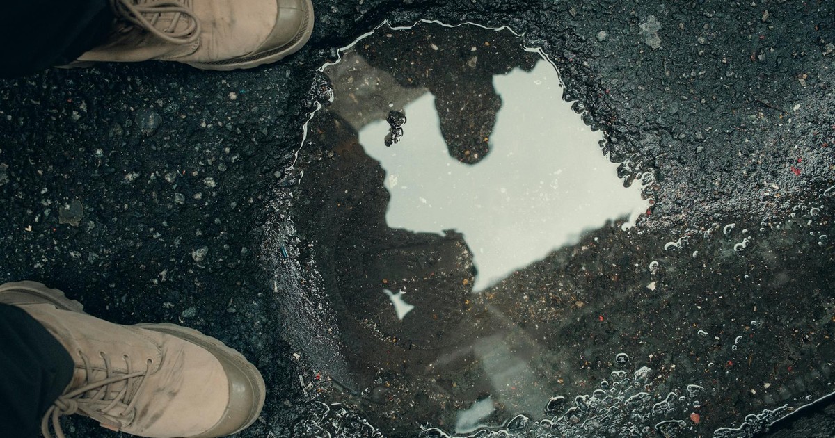 Close-up of shoes and a puddle on an asphalt surface reflecting people.
